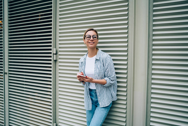 Fiatal vidám nő alkalmi ruhák és divatos szemüveg mosolyog és néz félre használata közben okostelefon és támaszkodik ablak vak az utcán - Fotó, kép