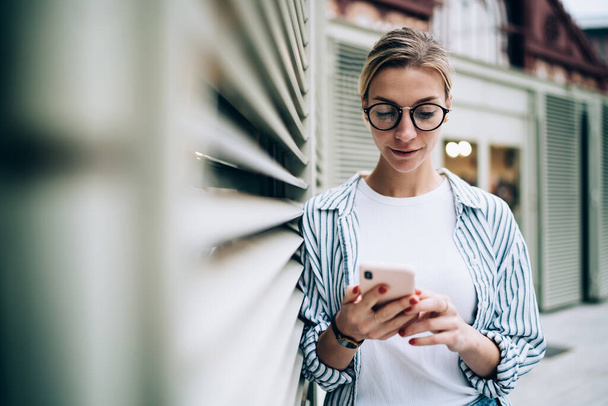 Young beautiful lady wearing casual clothes and trendy glasses using smartphone while leaning on building wall and reading latest news - Фото, зображення