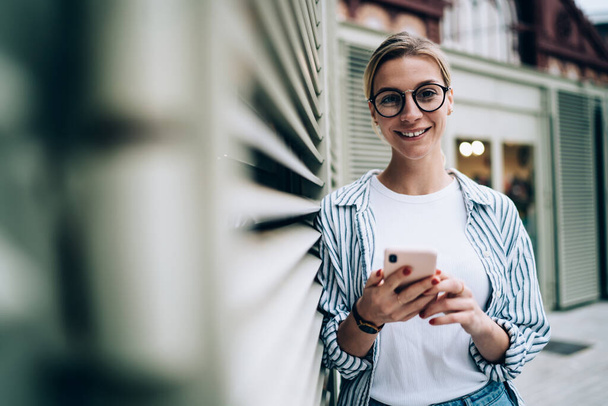 Cheerful blonde lady wearing casual clothes and trendy glasses smiling and looking at camera while leaning on wall and browsing smartphone - Фото, изображение