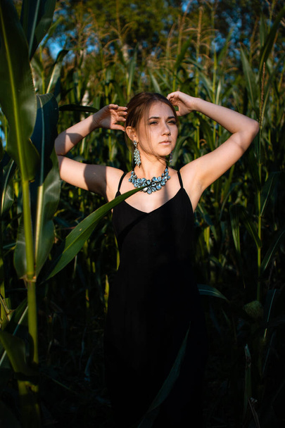Close-up of young woman in black dress, necklace and earrings between green leaves in a corn field in summer. Jewelry fashion style photo session. - Foto, afbeelding