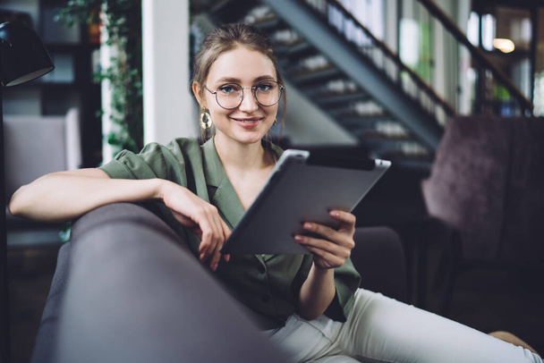 Adult modern casual female in round glasses holding tablet while sitting on sofa in library and smiling at camera - Φωτογραφία, εικόνα