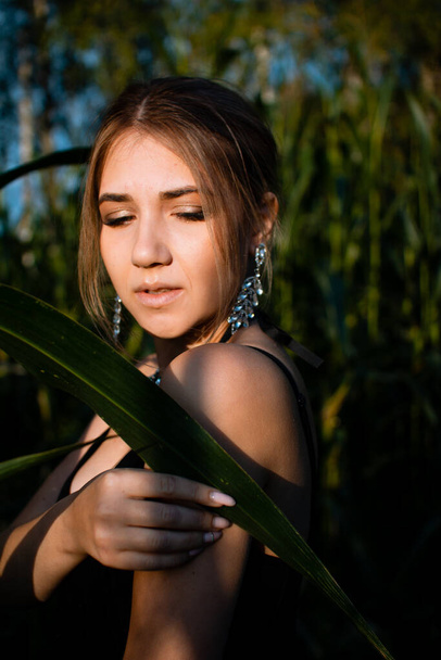 Close-up of young woman in black dress, necklace and earrings between green leaves in a corn field in summer. Jewelry fashion style photo session. - Zdjęcie, obraz