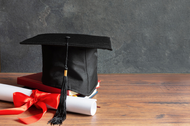 Academic cap and graduation diploma scroll and tied with red ribbon on wooden table over black chalkboard. - Photo, Image