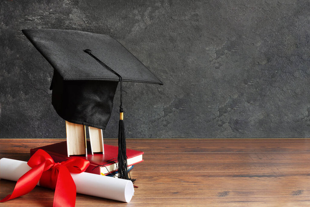 Tapa académica y diploma de graduación desplazarse y atado con cinta roja en la mesa de madera sobre pizarra negra
. - Foto, imagen