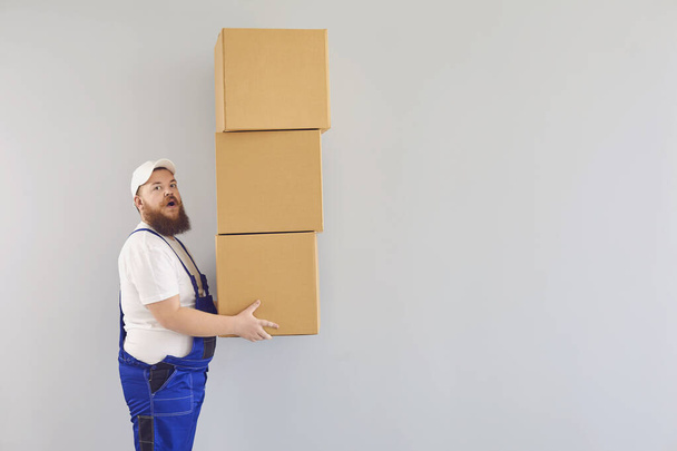 Funny fat bearded delivery courier loader man in a blue uniform with a cardboard box on a gray background. - Fotó, kép