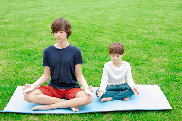 A young man with long hair does yoga outdoors. A teenager sits in a lotus position on the green grass. Copy space. Place for text. - Foto, afbeelding