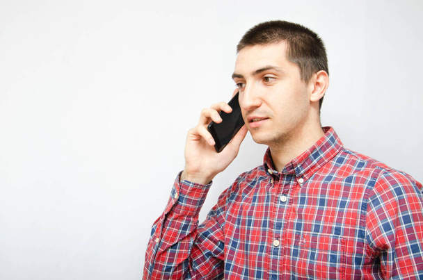 Young man in a plaid shirt with a smartphone on a white background. The guy holds a smartphone by the ear, talking on the phone. Anger, anger, surprise, attentiveness during a telephone conversation - Photo, Image