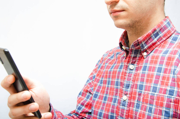 Young man in a plaid shirt with a smartphone on a white background. The guy looks at the smartphone and holds it in front of him - 写真・画像