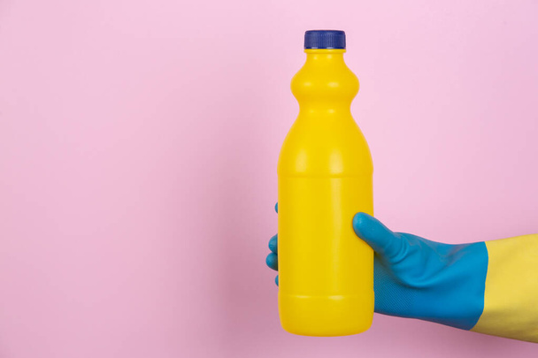 Рука в чистящих перчатках с бутылкой отбеливателя на розовом фоне
 - Фото, изображение