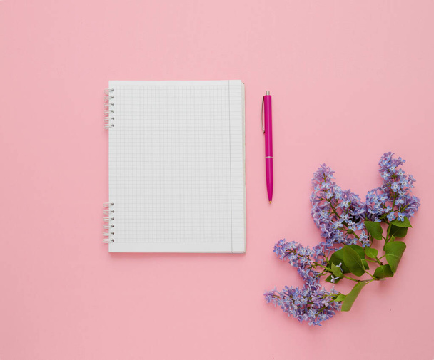 Top view of notebook, pen and lilac flower on a pink background. - Фото, изображение