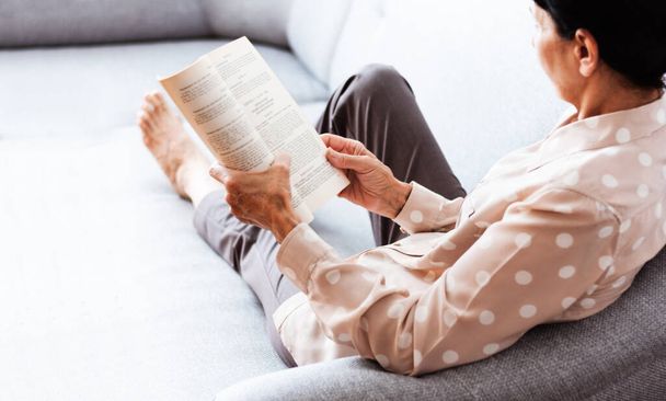Middle-aged brunette woman on the gray sofa reading book, soft focus, stay at home concept, cozy background - Foto, Bild