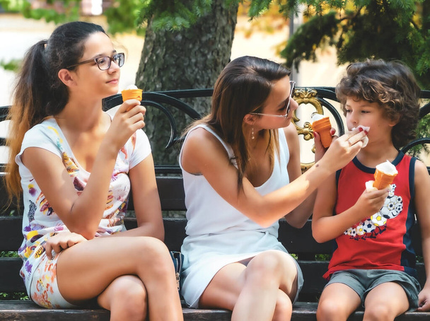 Happy family eating ice cream in a summer park. Sisters and little brother have fun. Walk in the green park. Mom clean mouth baby. - Zdjęcie, obraz