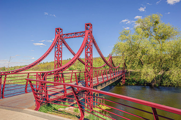 красивый железный красный подвесный пешеходный мост через реку Ирпин от набережной до зеленого леса
. - Фото, изображение