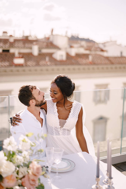 Úticél: képzőművészeti esküvő Firenzében, Olaszországban. Többnemzetiségű házaspár. Afrikai-amerikai menyasszony és kaukázusi vőlegény ülnek a tetőtéri esküvői asztalnál, kilátással a városra.. - Fotó, kép