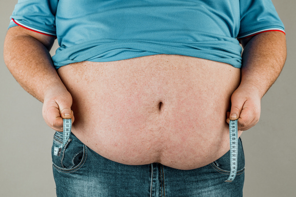 Un hombre mide su vientre gordo con una cinta métrica. sobre un fondo gris
 - Foto, Imagen