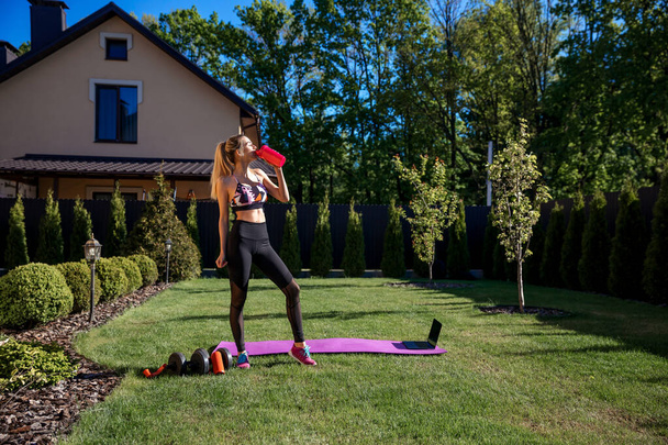 Pretty sportive fitness coach woman drinking water before online workout  training at the garden with fresh green grass near house.   - Photo, Image