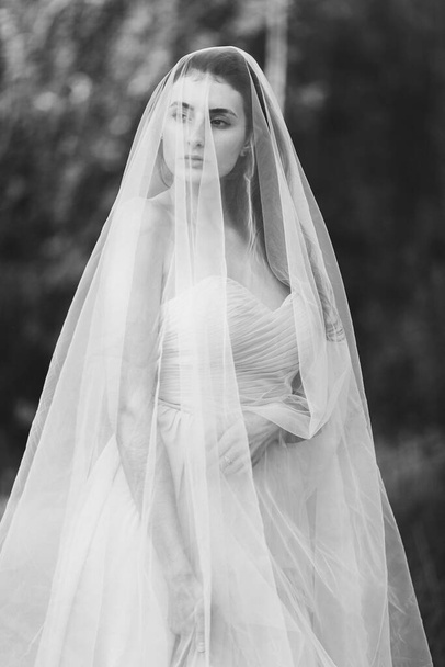 Portrait of a beautiful young brunette bride woman in a dress and with a long veil Curling in the wind on the nature garden in the summer. Fine art, wedding. - Fotó, kép