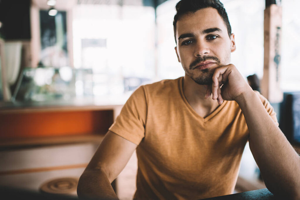 Optimistic handsome young male with mustache in casual wear leaning on hand taking break spending time in cafe looking at camera - Photo, Image