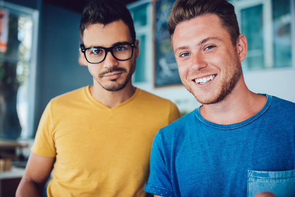 Cheerful smiling young guy in casual clothes spending time with calm handsome friend in glasses in cafeteria looking at camera - 写真・画像