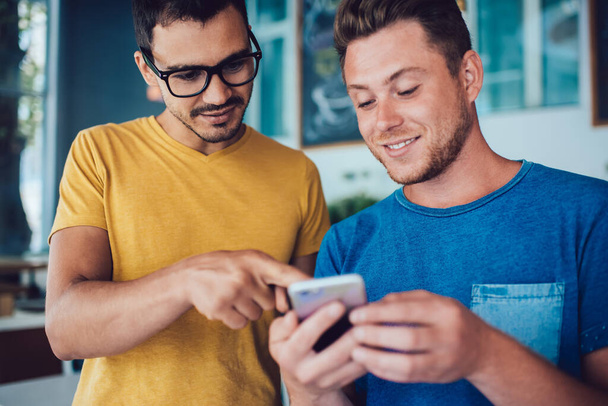 Joyful young man in casual clothes relaxing with content handsome friend in glasses pointing at screen of smartphone in cafeteria - Фото, изображение