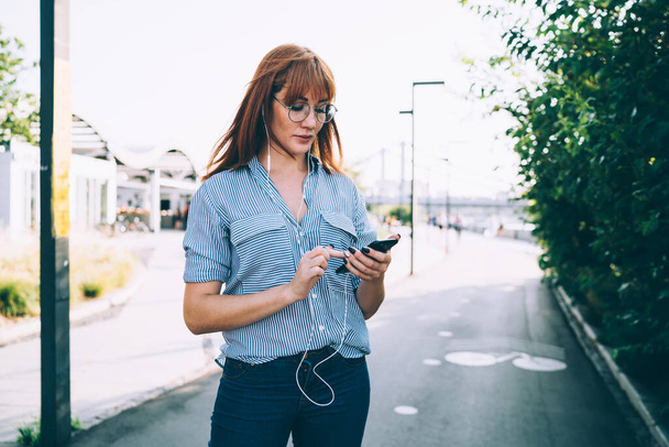 Young female in casual clothes using mobile phone and earphones and choosing playlist for listening while standing on road in city  - Фото, изображение