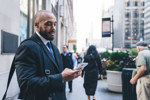 Handsome bald and bearded African American businessman in formal clothes standing and texting message on downtown street on blurred background - Фото, изображение