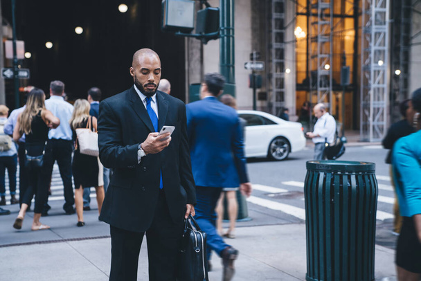 Confident bald African American bearded businessman in formal wear with leather briefcase using smartphone while walking on downtown street on blurred background  - Foto, afbeelding