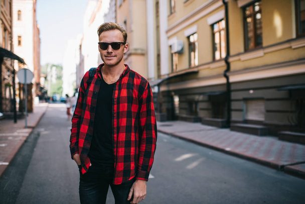 Handsome confident guy in sunglasses and checkered shirt with hand in pocket against blurred street and facades of multistory buildings in downtown - Foto, Imagem