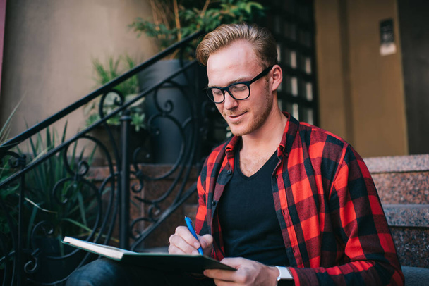 Cheerful content red haired male in glasses wearing checked red and black shirt making notes in notebook with pen sitting on granite stairs in city in summer  - Φωτογραφία, εικόνα