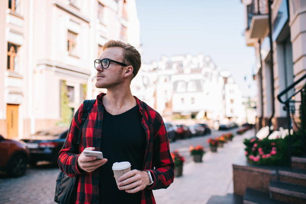 Serious male in casual wear and glasses with backpack taking cup of coffee surfing mobile walking in street of city - Photo, Image