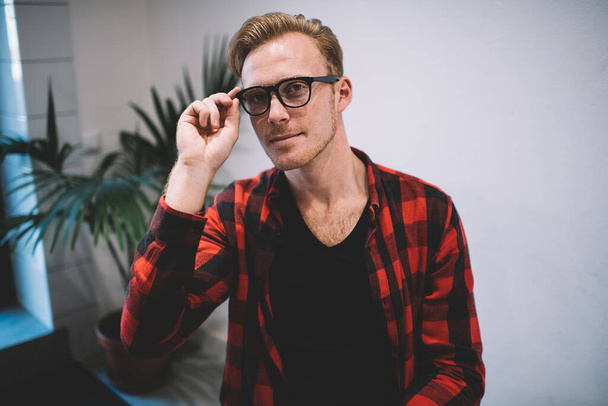 Portrait of handsome young man with blond hair in casual clothes adjusting black glasses relaxing in cafeteria looking at camera - Foto, immagini