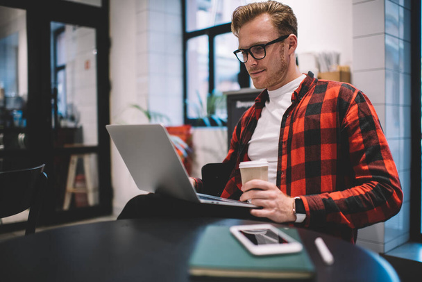 Immersed smiling clever freelancer in checkered shirt browsing laptop on knees while working remotely with coffee takeaway in modern cafeteria - 写真・画像