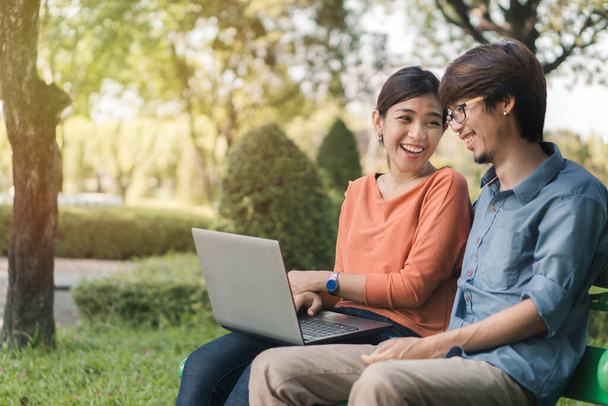 Young business asian man and woman working with them laptop and look at the face and smile together on a bench in the park outdoors on vacation time, Selective focus. - 写真・画像