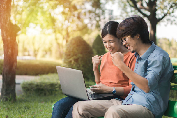 Couple young asian man and woman working with laptop and enjoying cheerful his online winner success  on a bench in the park outdoors on vacation time. - Photo, Image