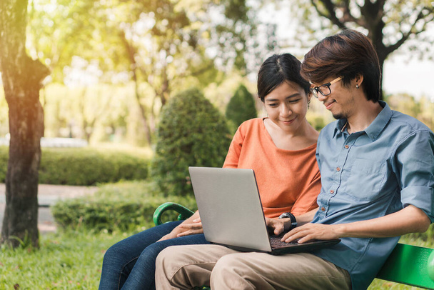 Young asian man and woman working with them laptop on a bench in the park outdoors on vacation time. - Zdjęcie, obraz