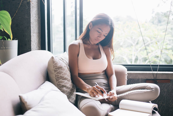 Smart slim Asian woman in sportswear reading book and writing in notebook sitting on couch with pillows against window in summer day - Photo, Image