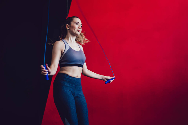 Positive smiling young lady in sport clothes using jump rope skipping doing fitness exercise training legs spending time in gym - Foto, Imagem