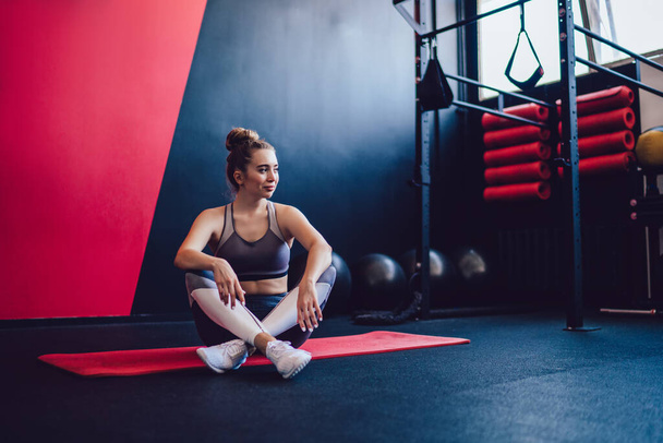 Smiling relaxed young female athlete sitting with crossed legs on mat near sports equipment during break in gym looking away - 写真・画像