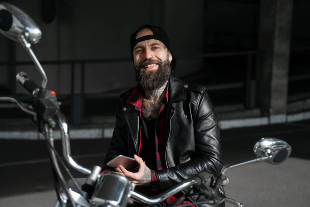 El viajero tiene un viaje en bicicleta, sonriendo brutal barbudo motorista hombre de negro con teléfono celular móvil usando mapa en línea en bicicleta, estilo de motocicleta, hombre elegante, estilo de vida de la gente
.  - Foto, imagen