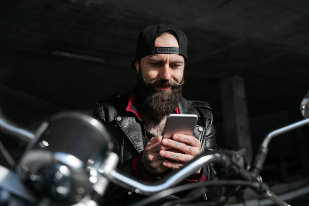 Brutale biker uomo barbuto in nero con telefono in bicicletta, stile moto, uomo alla moda utilizzando il cellulare e le tecnologie Internet online, comunicazione tramite connessione 5g. Stile di vita
  - Foto, immagini