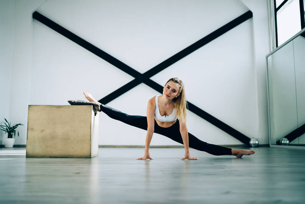 Magnificent slim determined woman in sport clothes smiling at camera while doing front split exercise leaning on wooden box and floor against black sports wall resistance bands in cozy modern gym - Foto, imagen
