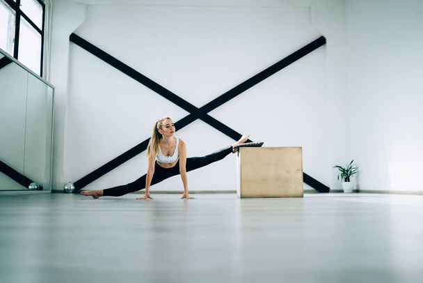 Calm slim adult athlete in sport clothes looking away and working out leg muscles while doing front split exercise leaning on wooden box and floor against black sports wall resistance equipment in cozy modern sport club - Φωτογραφία, εικόνα