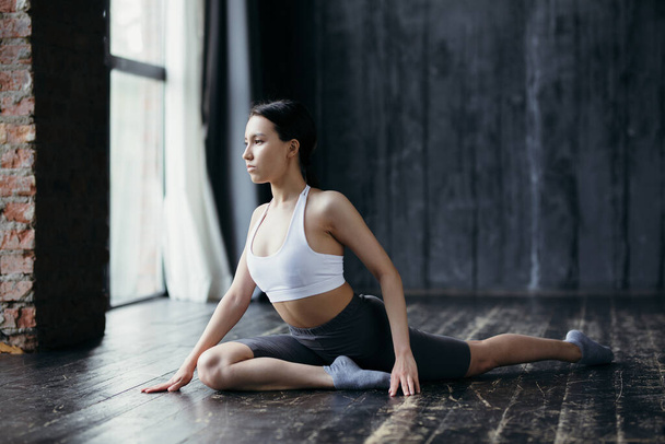 The athlete sits in a fitness studio with large windows on the floor and is engaged in stretching for longitudinal twine - 写真・画像