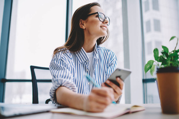 Happy young female in spectacles smiling and looking away while suing smartphone and writing in planner during work in modern workplace - Фото, изображение