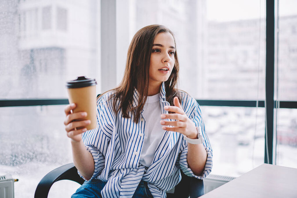 Young woman with cup of drink to go gesticulating and telling story while sitting against window and resting during break in modern workspace - Фото, изображение