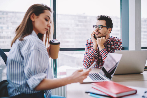 Pensive male office employee in stylish casual wear calling on smartphone while working with young female coworker at modern office  - Photo, image