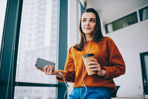 Pleased youthful charming woman in casual clothes and earbuds listening music on smartphone and holding takeaway cup while standing during coffee break in office - Photo, Image