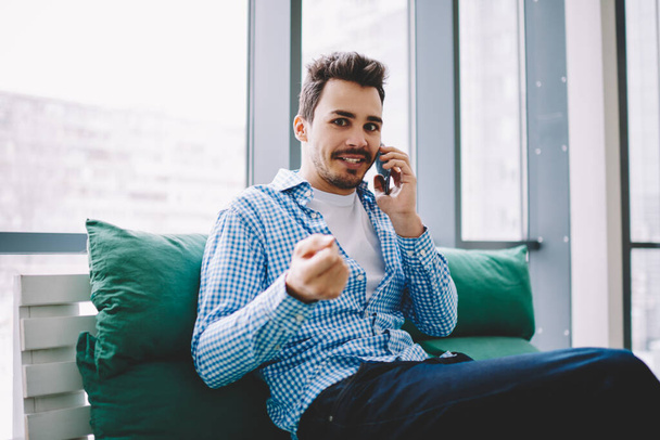 Content surprised guy in casual wear with fist up using phone chatting with coworker celebrating success sitting at workplace looking at camera - Foto, Bild