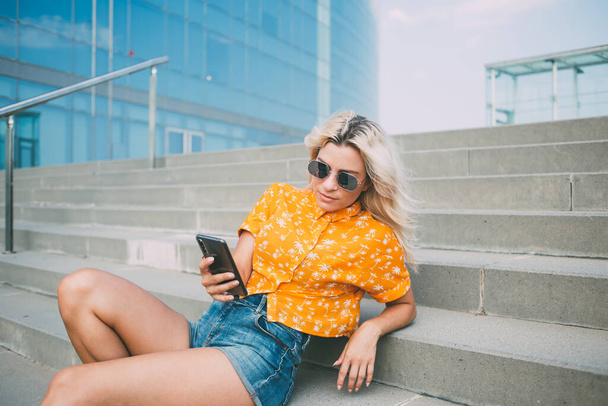Relaxed young lady in stylish outfit and sunglasses lying on steps and resting while spending time on street of modern city - Фото, изображение
