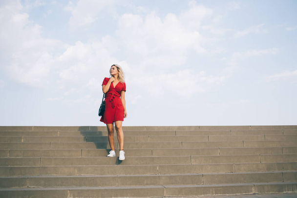 Young fair haired woman in short red dress standing on large stone staircase while looking away on background with sky - Foto, Imagen